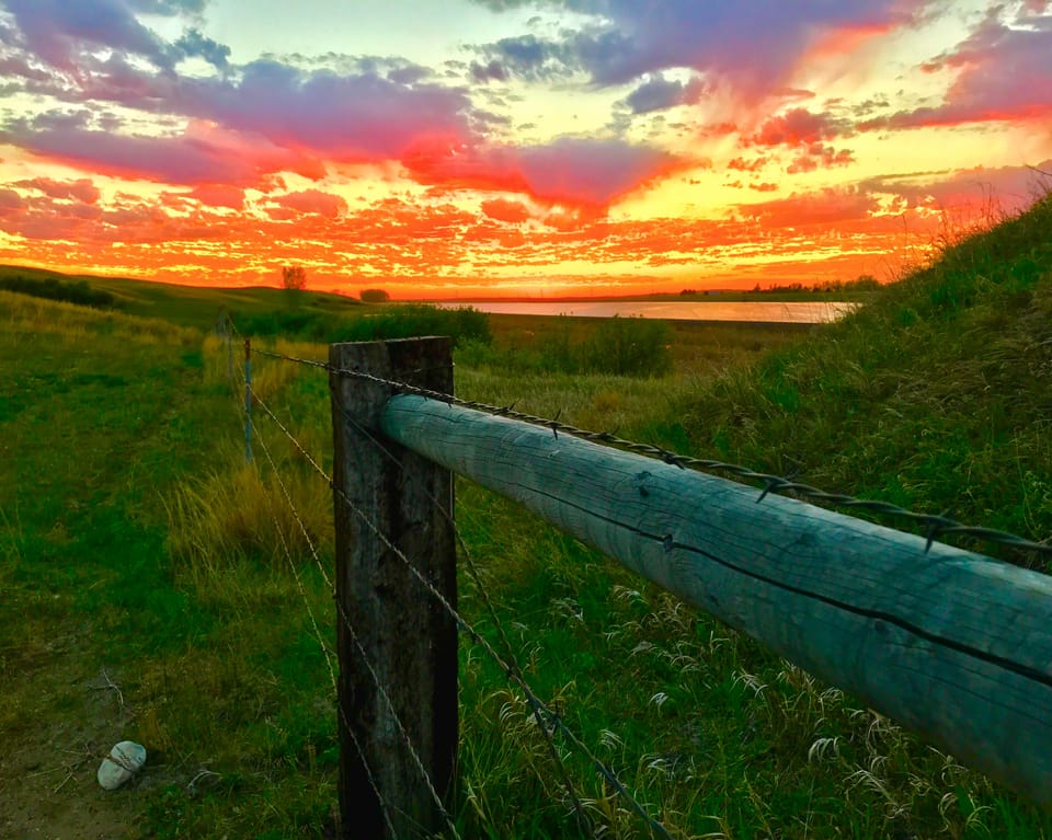 A weathered post and barbed wire in grass, with water and a red evening sky beyond.