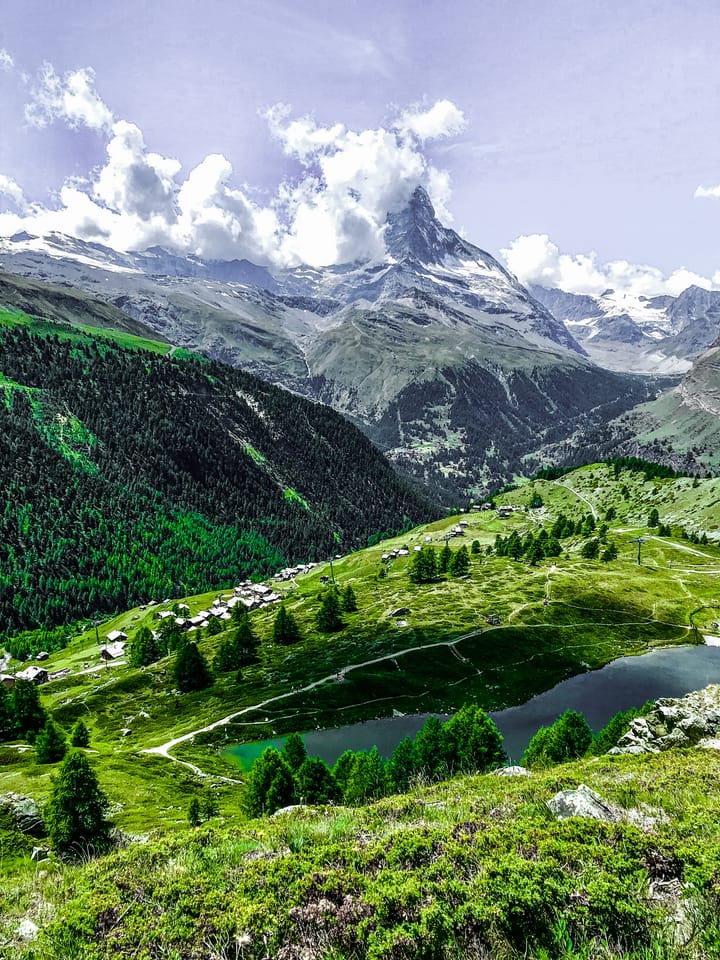 A high green valley with a lake, a village on the slope, and a pointed snow peak.