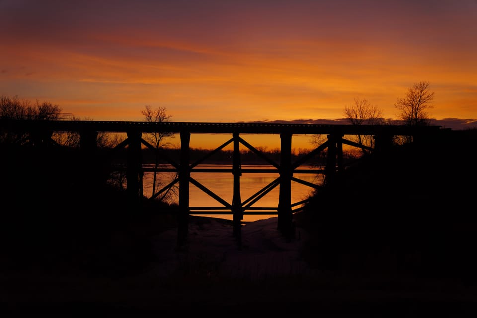 A wooden trestle in black silhouette against an orange and violet dusk, with water under the span.