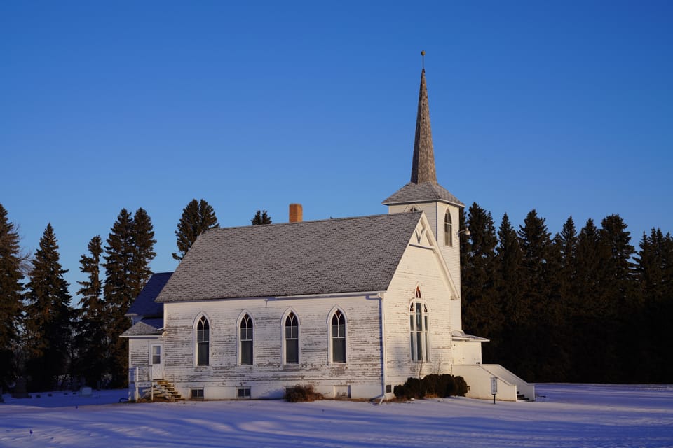 A white country church with a steeple, Gothic windows, snow, and a line of spruce.