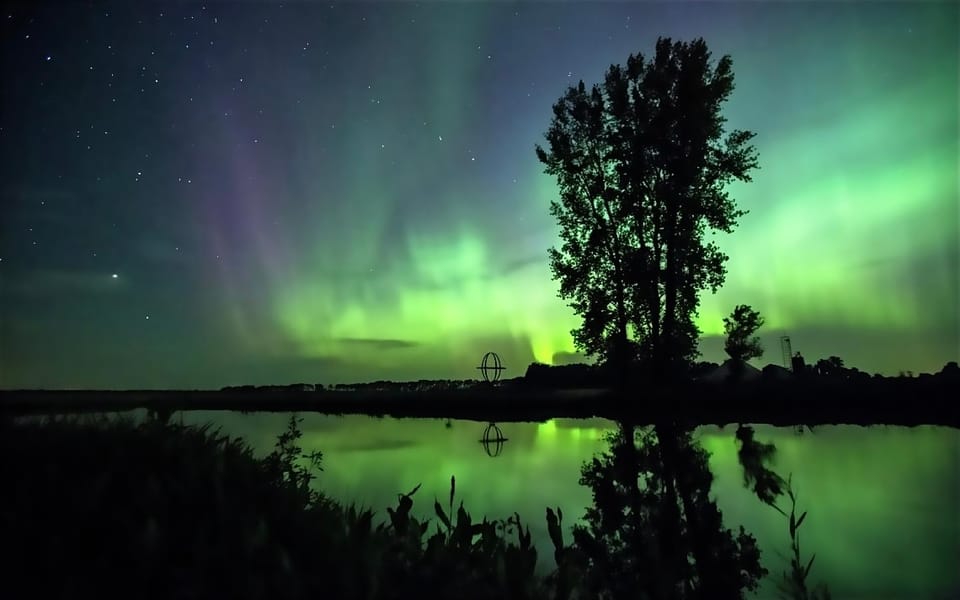 Green aurora over still water, a silhouetted tree, and a distant structure.