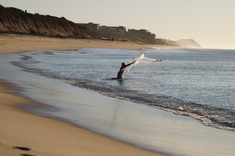 A fisherman casts a net in the evening shallows at Cabo San Lucas, Mexico, with cliffs and a long beach behind him.